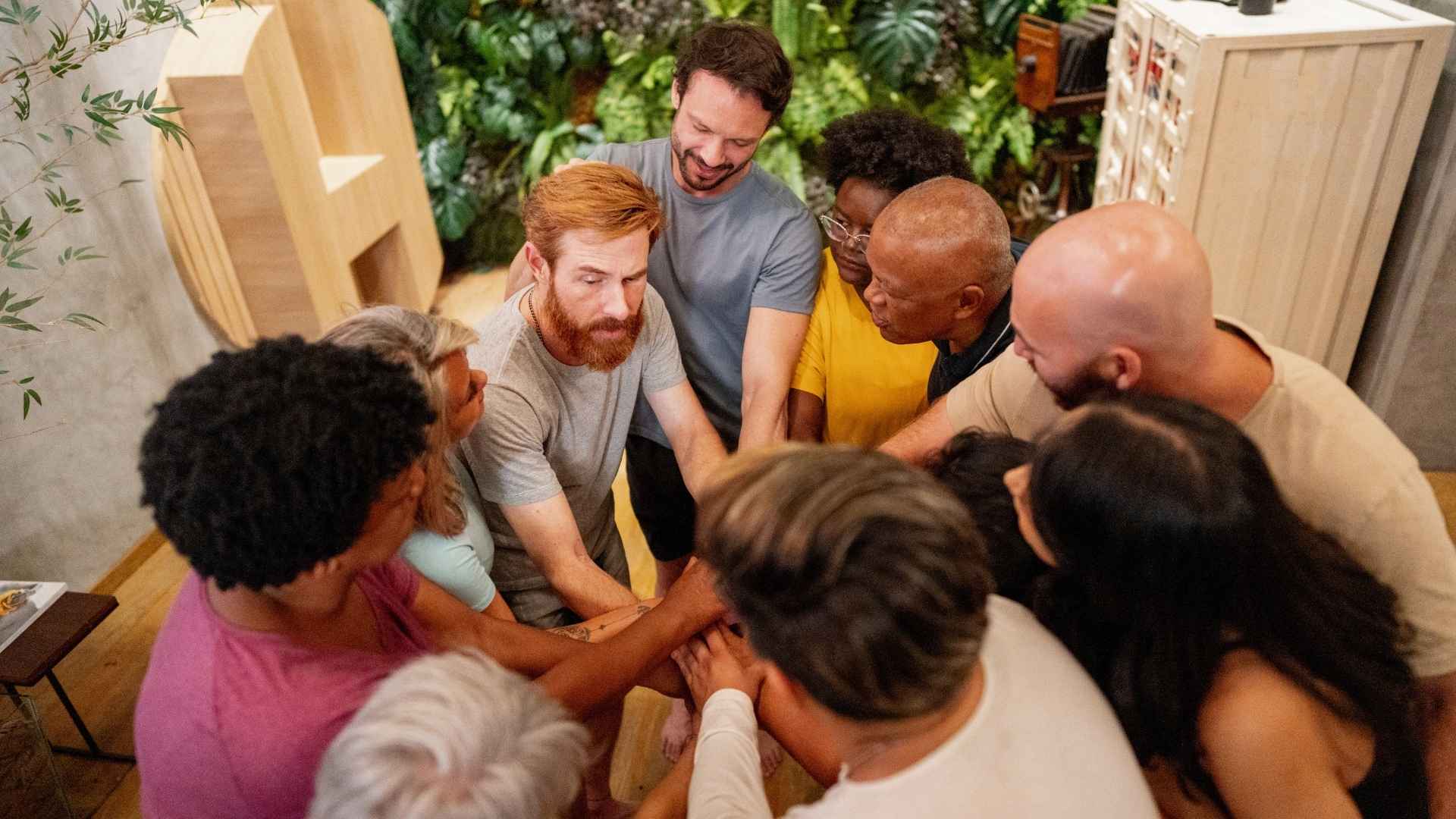 people gathered in a room forming a circle with hands in center with a plant wall behind